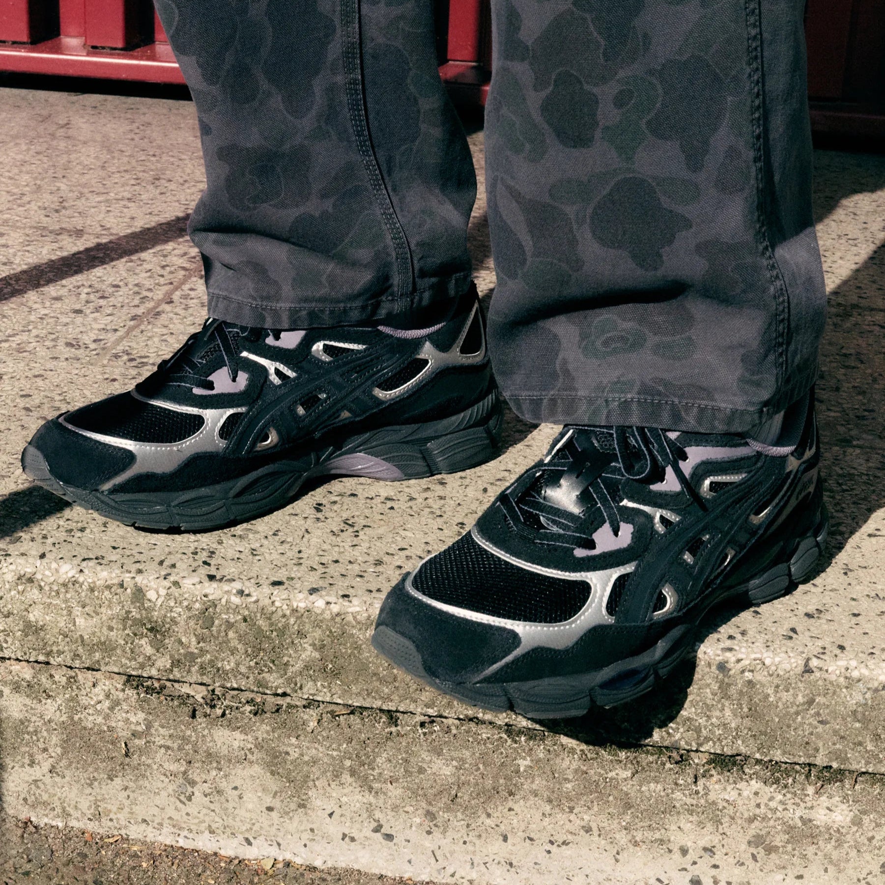 Black and gray sneakers worn with camouflage pants on a concrete surface.